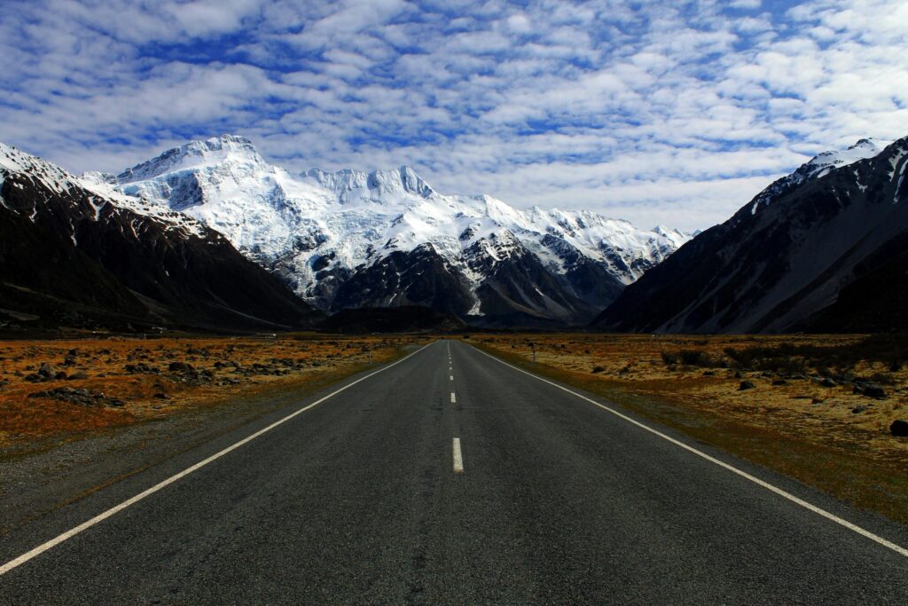 A deserted road leading to majestic snow-covered mountains under a cloudy blue sky.