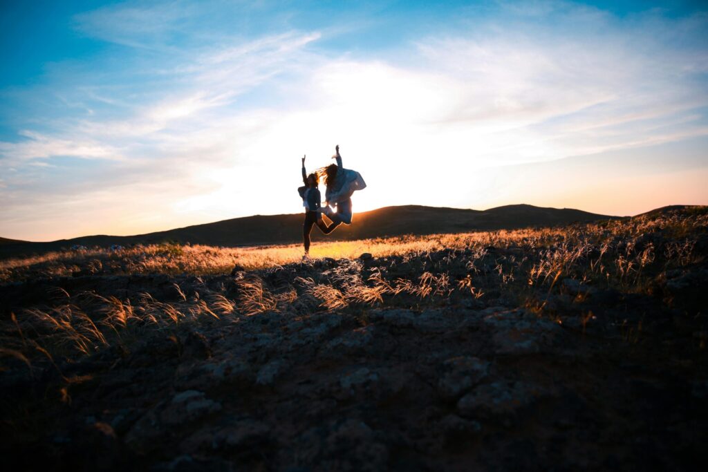 Two friends jump joyfully at sunset in a rocky field, creating a silhouette against the vibrant sky.