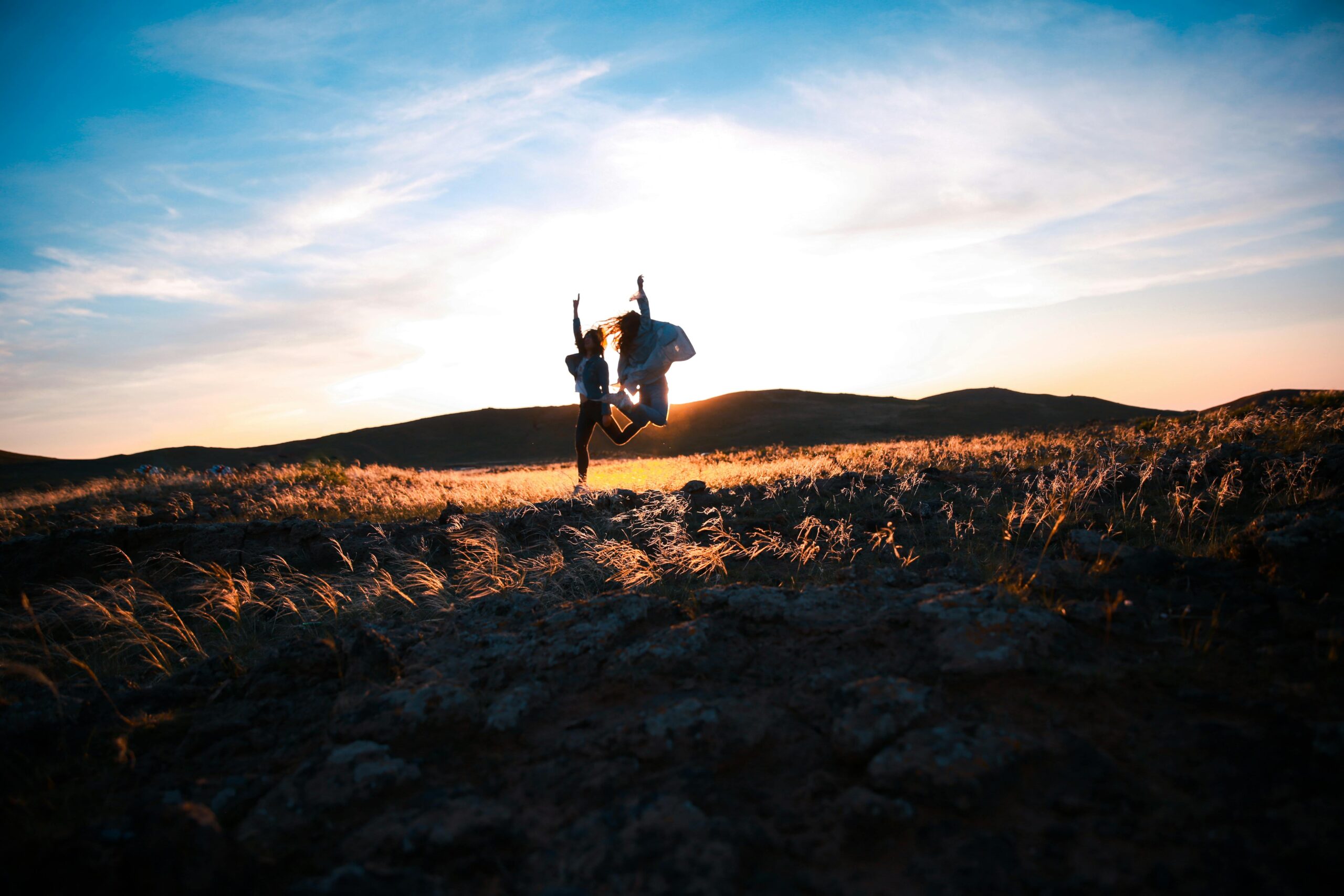 Two friends jump joyfully at sunset in a rocky field, creating a silhouette against the vibrant sky.